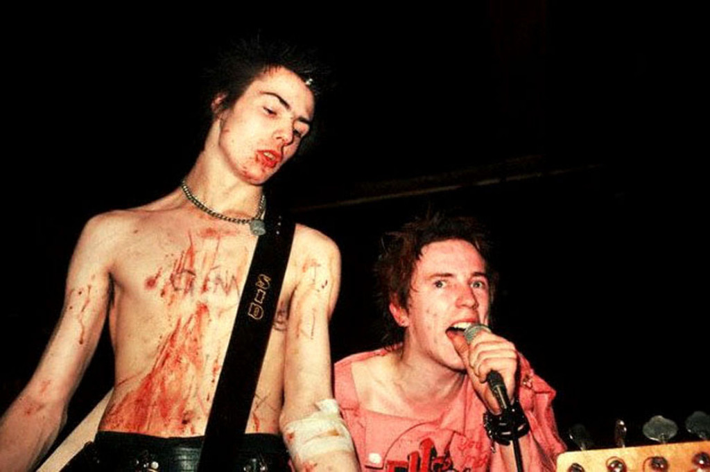 ca. 1977-1978 --- Sid Vicious, bassist, and Johnny Rotten, singer, for the punk band the Sex Pistols, performing a concert. Rotten crouches on the right, underneath Vicious's bass. Rotten wears a pink, obscene T-shirt. Vicious is shirtless. His chest is bleeding because he has recently carved the words, "Gimme a Fix" into it with a razor. Undated (1970s). --- Image by ¬© Lynn Goldsmith/Corbis