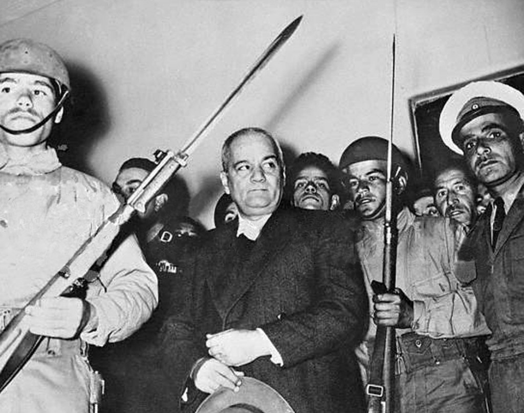 Original Caption: Jail Breaking Iran Red Leader Recaptured. Tehran, Iran: Dr. Moteza Yazdi, leader of the Communist Party of Iran, is shown surrounded by armed guards after his arrest while escorting to party girls recently. Yazdi had been in hiding since August, 1953. He was serving a prison term of 15 years following this trial with other Communist Party members when he broke jail. He now faces another trial and possible death.